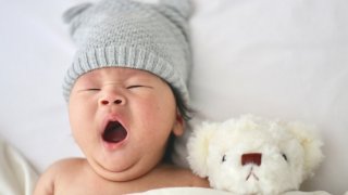 Baby yawning in bed next to a teddy bear