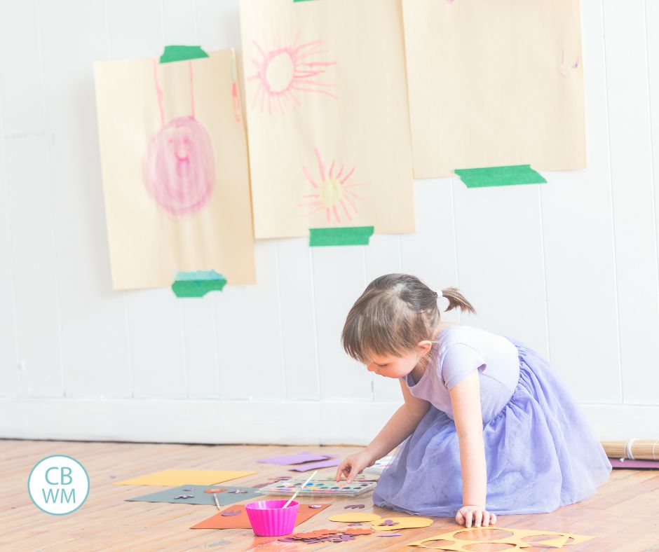 Girl doing art on the floor with her pictures hanging on the wall behind her