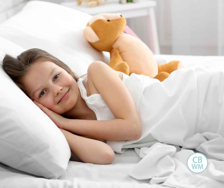 Child wearing white laying in a bed of white bedding smiling and looking toward the camera
