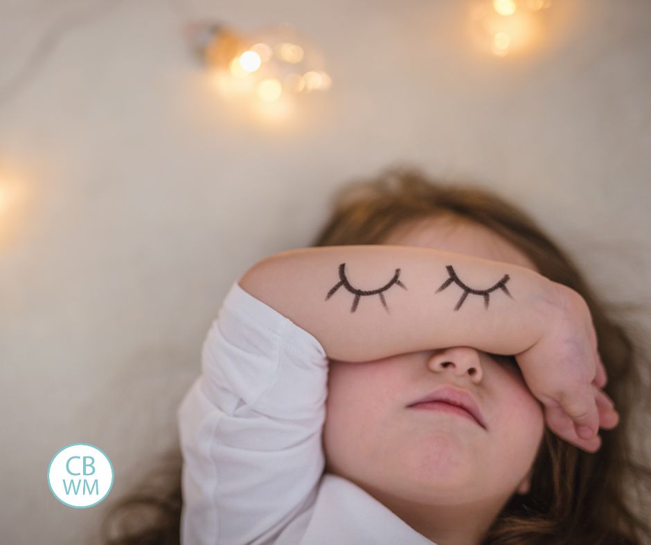 Preschooler lying down and covering her eyes with her forearm. Her arm has closed eyes drawn on it. 