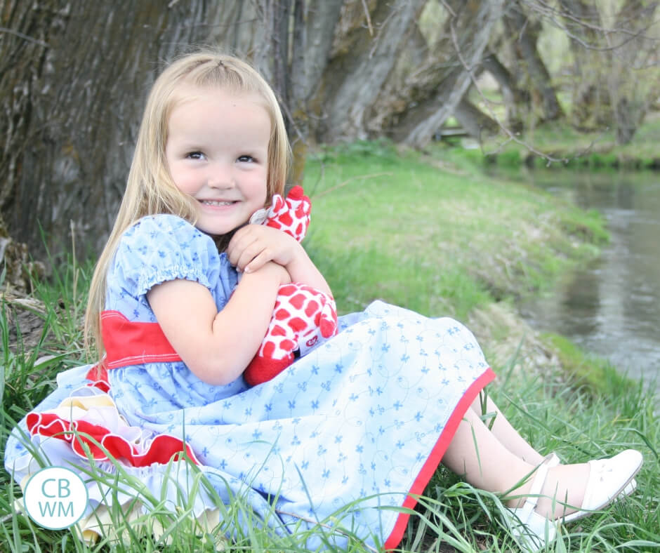 Preschool girl holding a stuffed animal as she sits on the bank of a creek