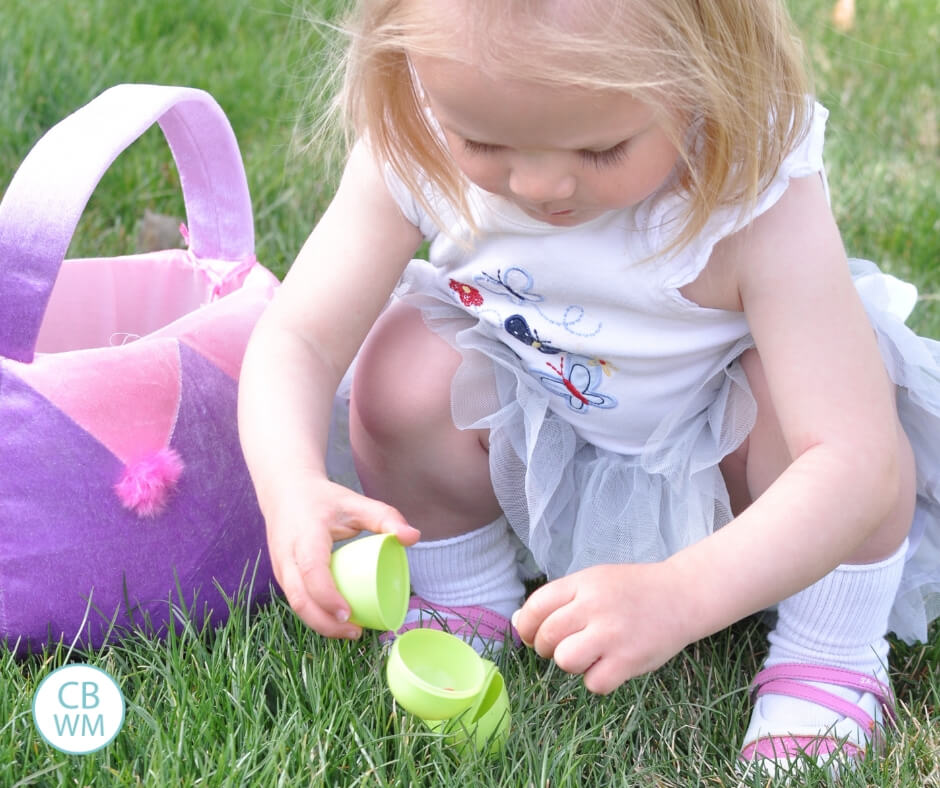 toddler opening Easter egg on the grass next to an Easter basket