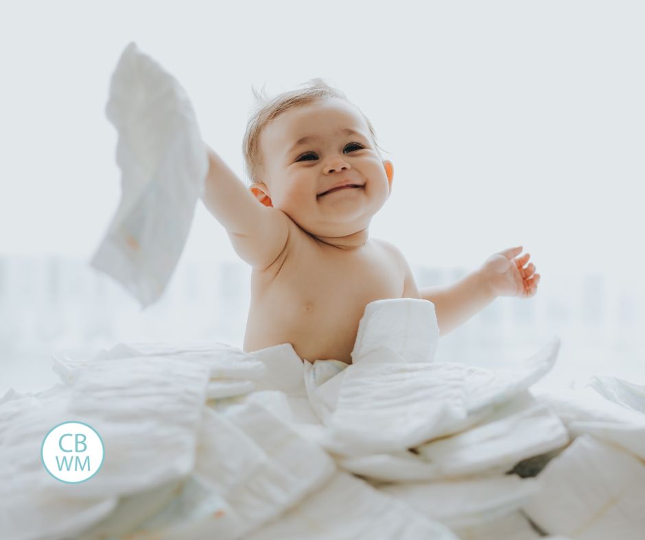 Baby sitting in pile of diapers, smiling and holding one diaper up