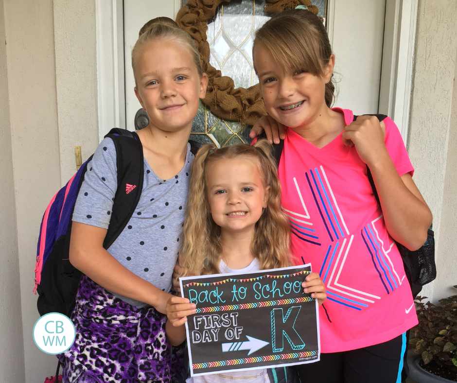 First day of Kindergarten for Brinley. She is standing between her two older sisters and holding a sign for her first day
